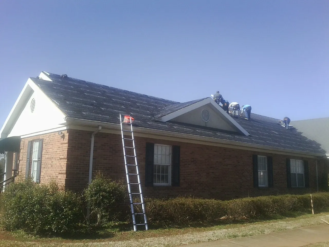 Crew tearing shingles off a brick ranch roof with a ladder set — Emergency Roof Repair work by our crew in Dunbar