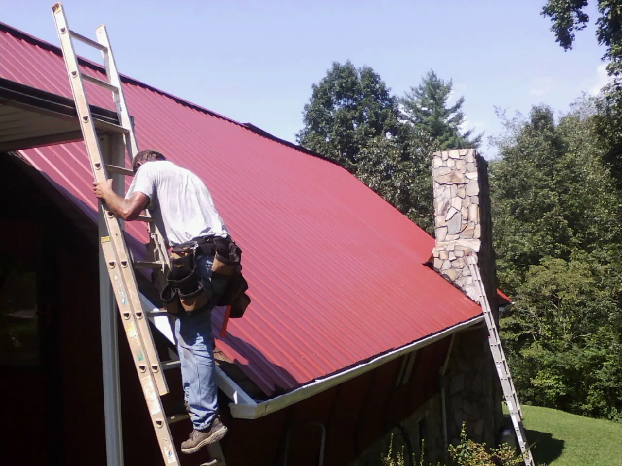 Roofer on a ladder beside a red metal roof and stone chimney — Soffit & Fascia Repair by Roofing Brothers in Dunbar